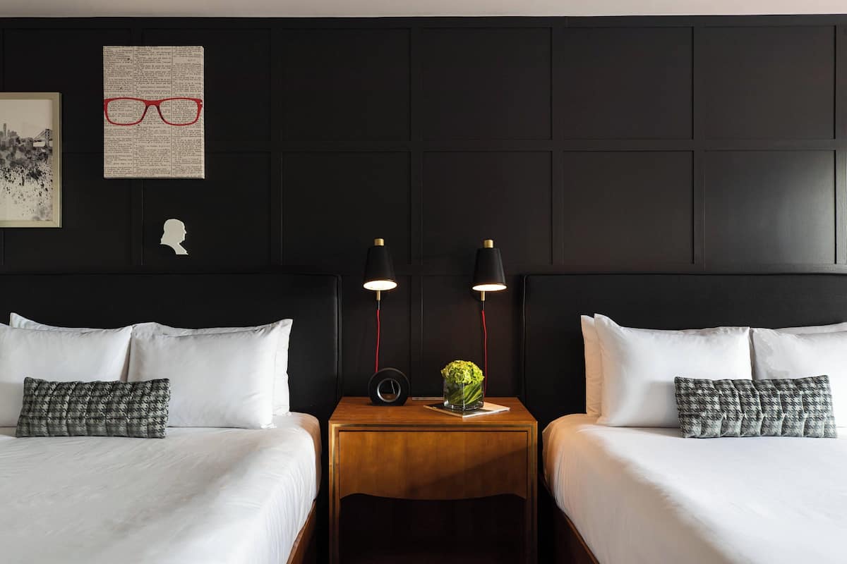 Une chambre d’hôtel avec deux lits, du linge de lit blanc, une table de chevet en bois ornée d’un vase de fleurs et des œuvres d’art mural, notamment une paire de lunettes et une silhouette.