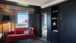 Ein modernes Hotelzimmer mit einem roten Sofa, einem Beistelltisch, einer Lampe, einem Kleiderschrank und Blick auf die Stadt.
