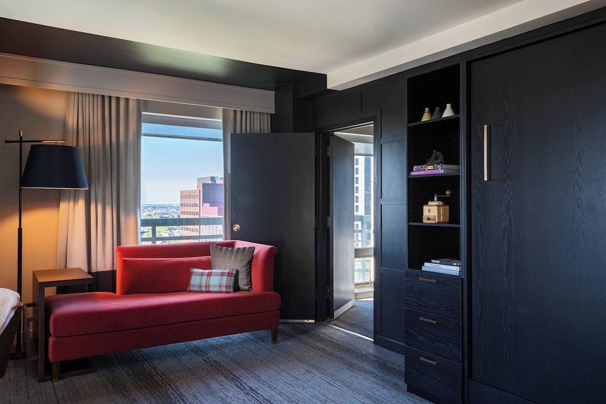 A modern hotel room with a red sofa, a side table, a lamp, a wardrobe, and a view of the city.