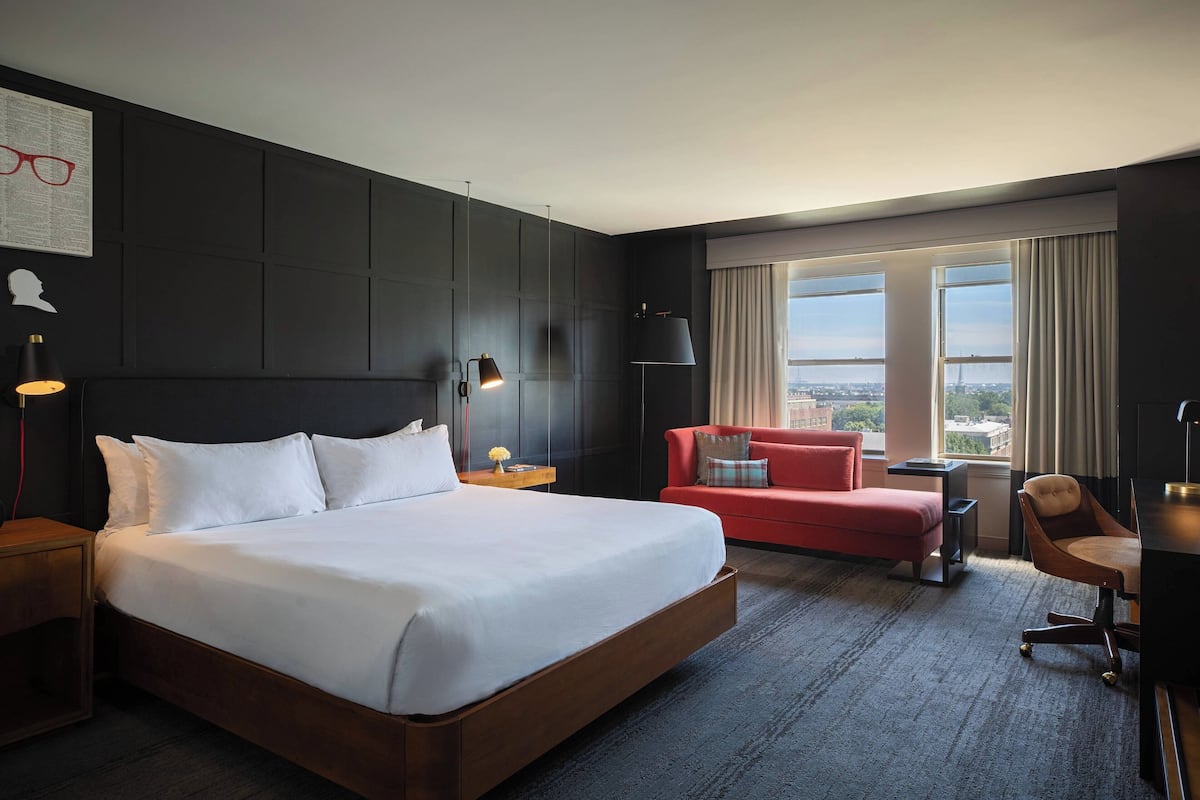Une chambre d’hôtel avec un grand lit, un canapé rouge, un bureau avec une chaise et une vue sur la ville grâce à de grandes fenêtres.