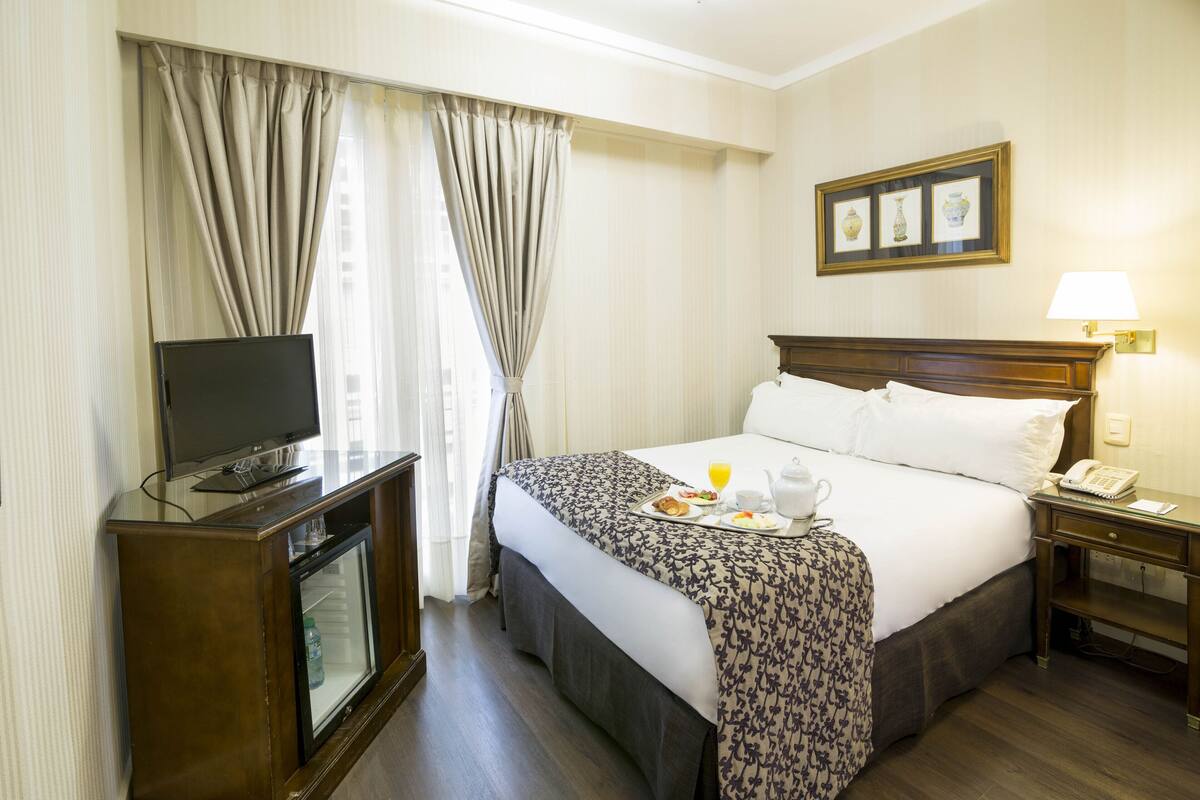 A hotel room with a bed, a television on a stand, a bedside table with a lamp, and a framed picture on the wall.