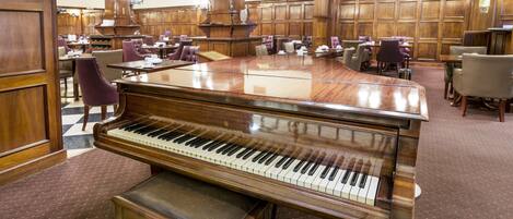 A grand piano in a spacious room with wooden paneling and multiple tables and chairs.