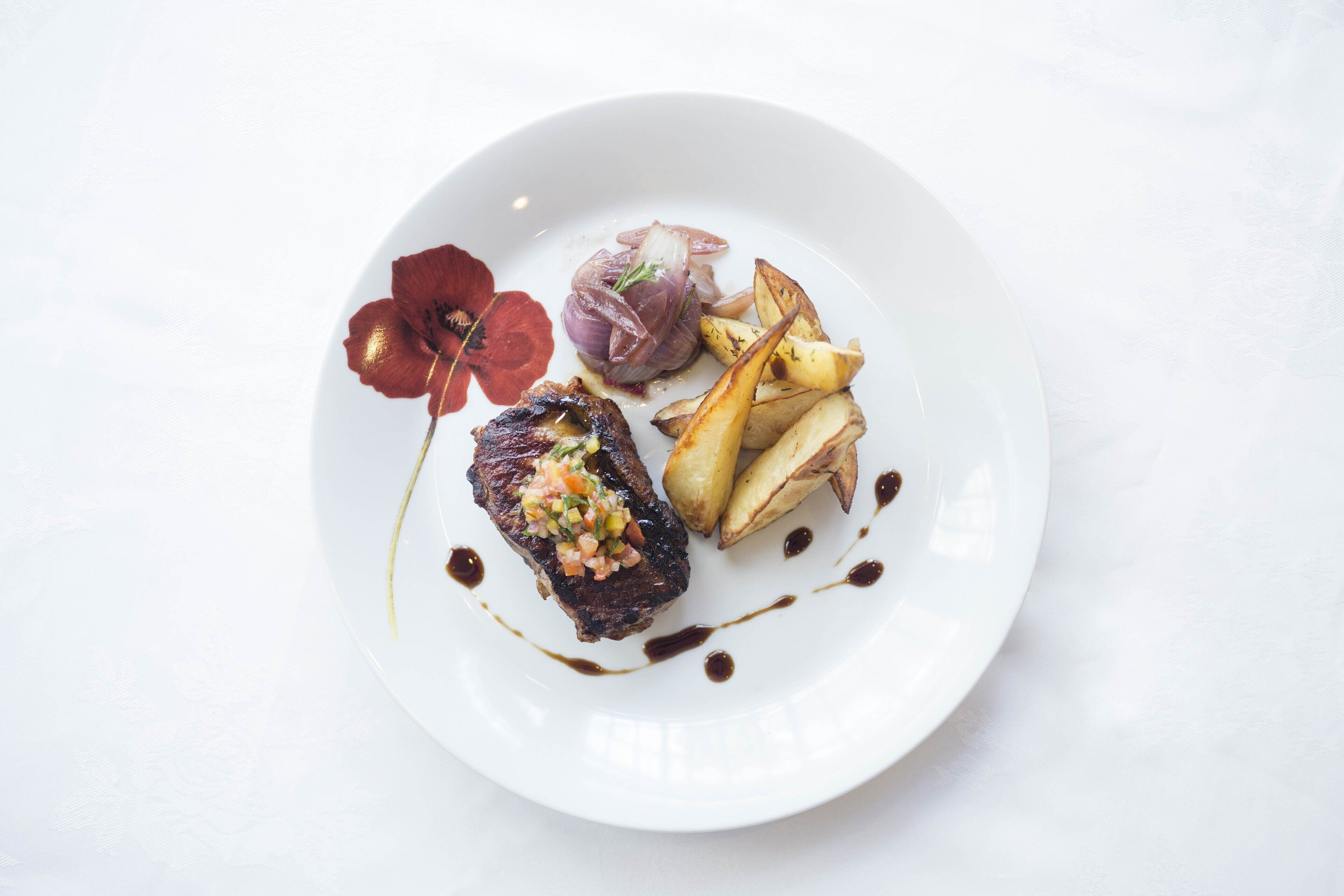 Une assiette blanche sur laquelle repose un steak, surmonté d’une salsa colorée, accompagné de pommes de terre rôties et décoré d’une fleur rouge.