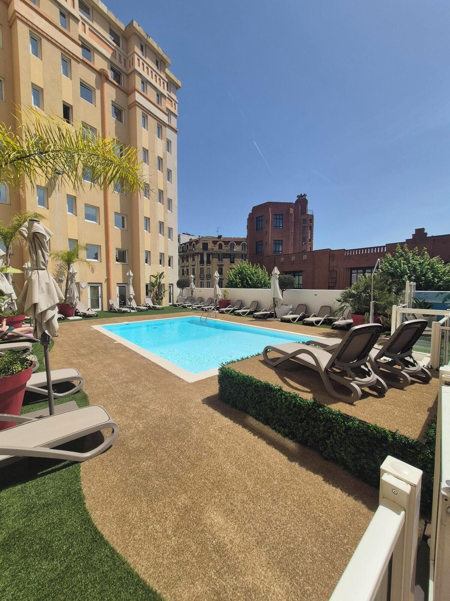 A rooftop pool area with lounge chairs and umbrellas, surrounded by buildings.