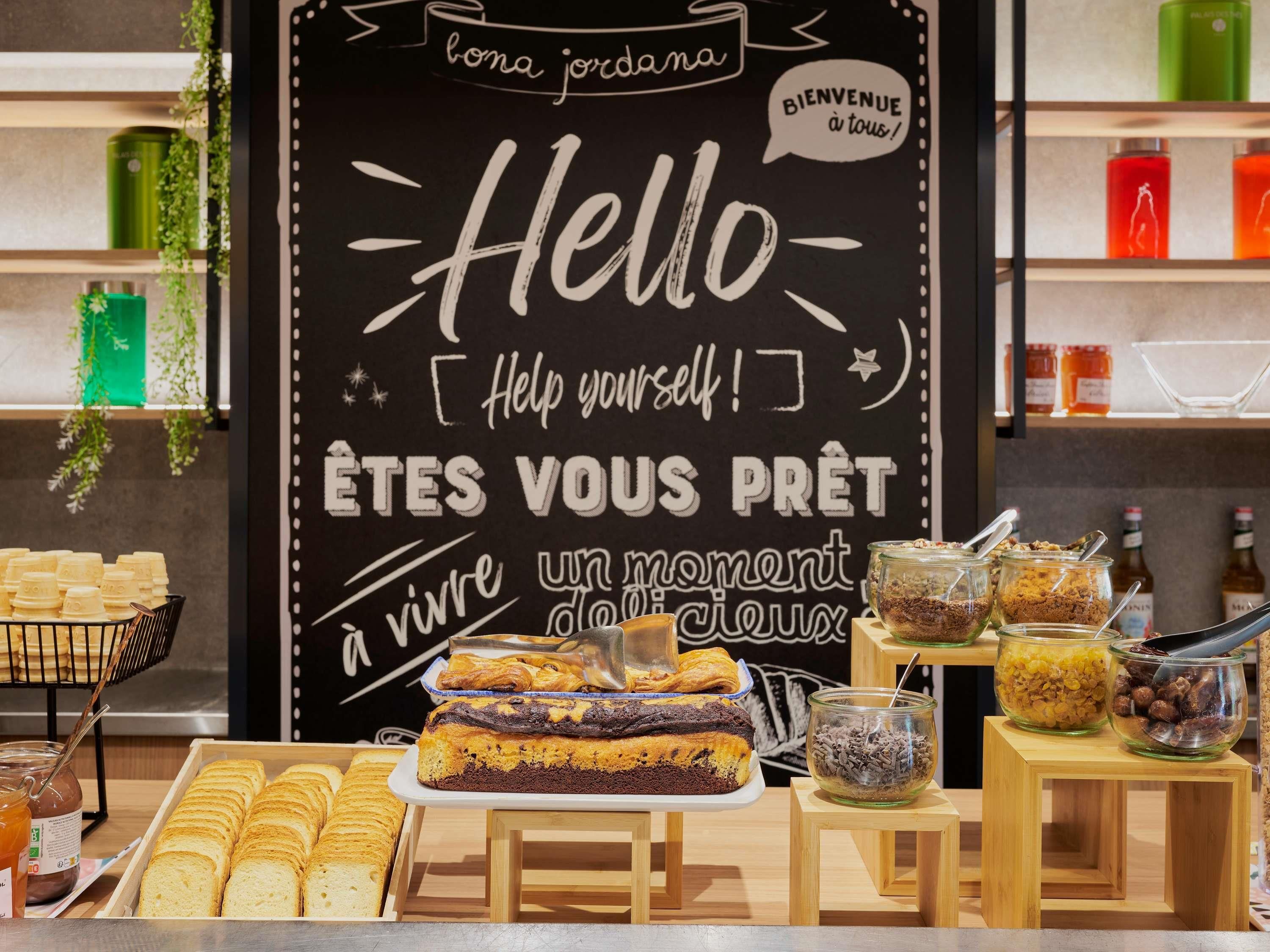 Een bakkerijdisplay met gebak, een schoolbord waarop staat 'Hallo [Help yourself!]' en 'ÊTES VOUS PRÊT à vivre un moment délicieux', en potten met spreads op houten standaardjes.
