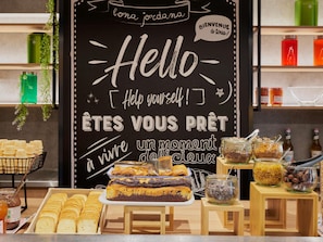 A bakery display with pastries, a chalkboard sign with 'Hello [Help yourself!]' and 'ÊTES VOUS PRÊT à vivre un moment délicieux', and jars of spreads on wooden stands.