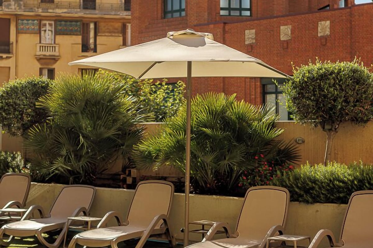 A pool area with lounge chairs and a large umbrella, surrounded by greenery and a red brick building in the background.