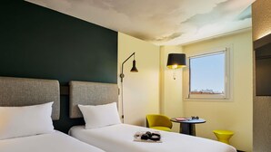 A hotel room with two beds, a small table with a chair, a lamp, and a window. - ibis Nice Centre Gare (Nice)