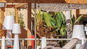 Un área de estar al aire libre iluminada por el sol, con sillones de descanso y un gran paraguas, rodeada de abundante vegetación y la fachada de un edificio.