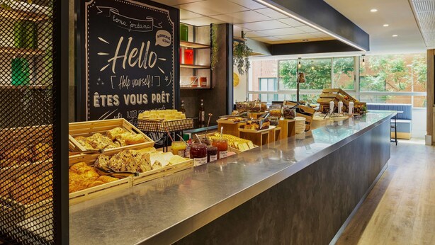 A breakfast buffet with various food items, a chalkboard with a greeting message, and a seating area with a view of the outdoors. - ibis Nice Centre Gare (Nice)