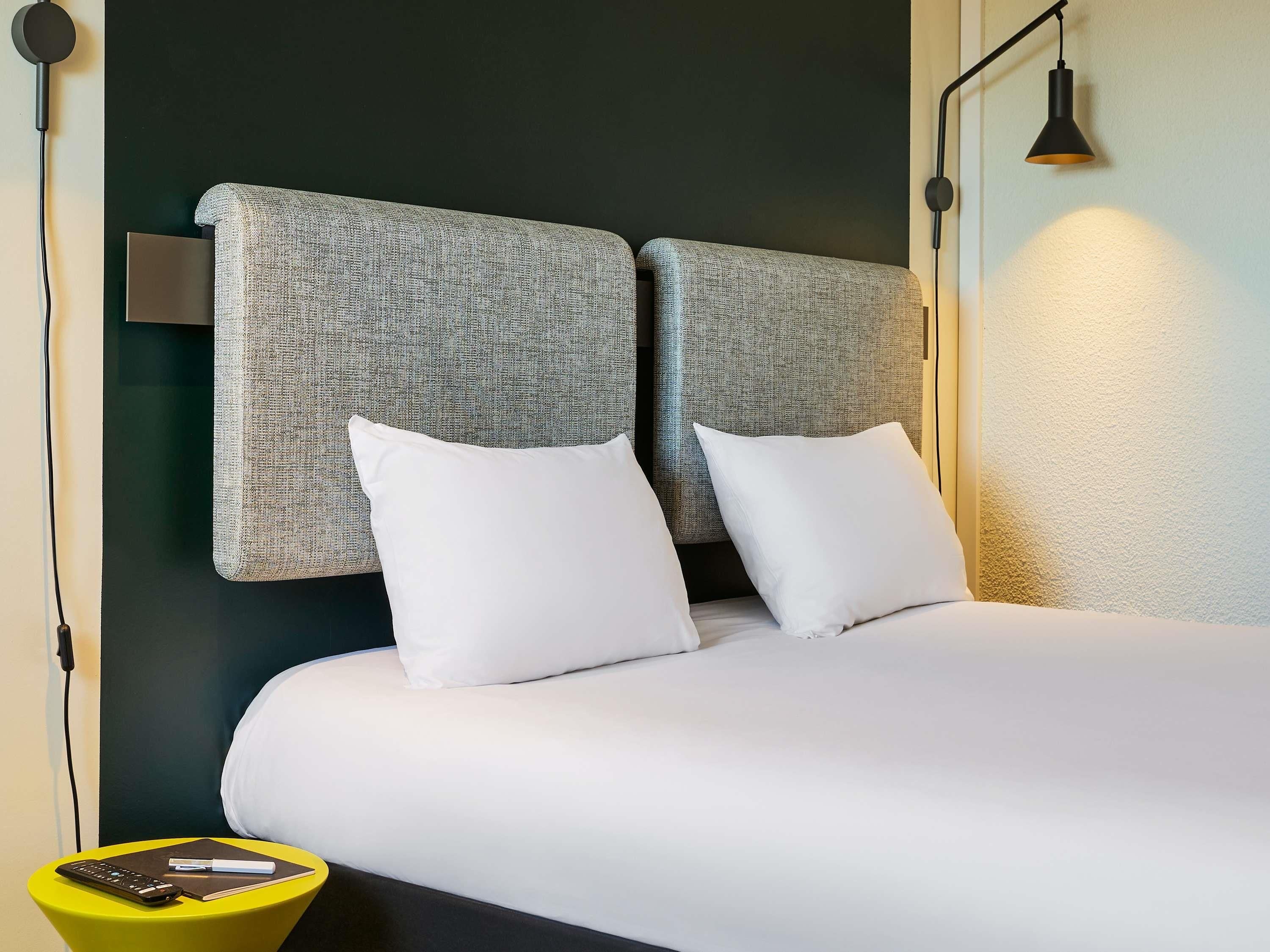Habitación de hotel con cama, dos almohadas, mesita de noche con un control remoto y una libreta, y una lámpara.