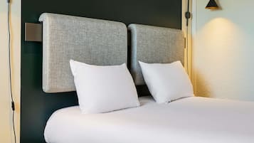 Habitación de hotel con cama, dos almohadas, mesita de noche con un control remoto y una libreta, y una lámpara.