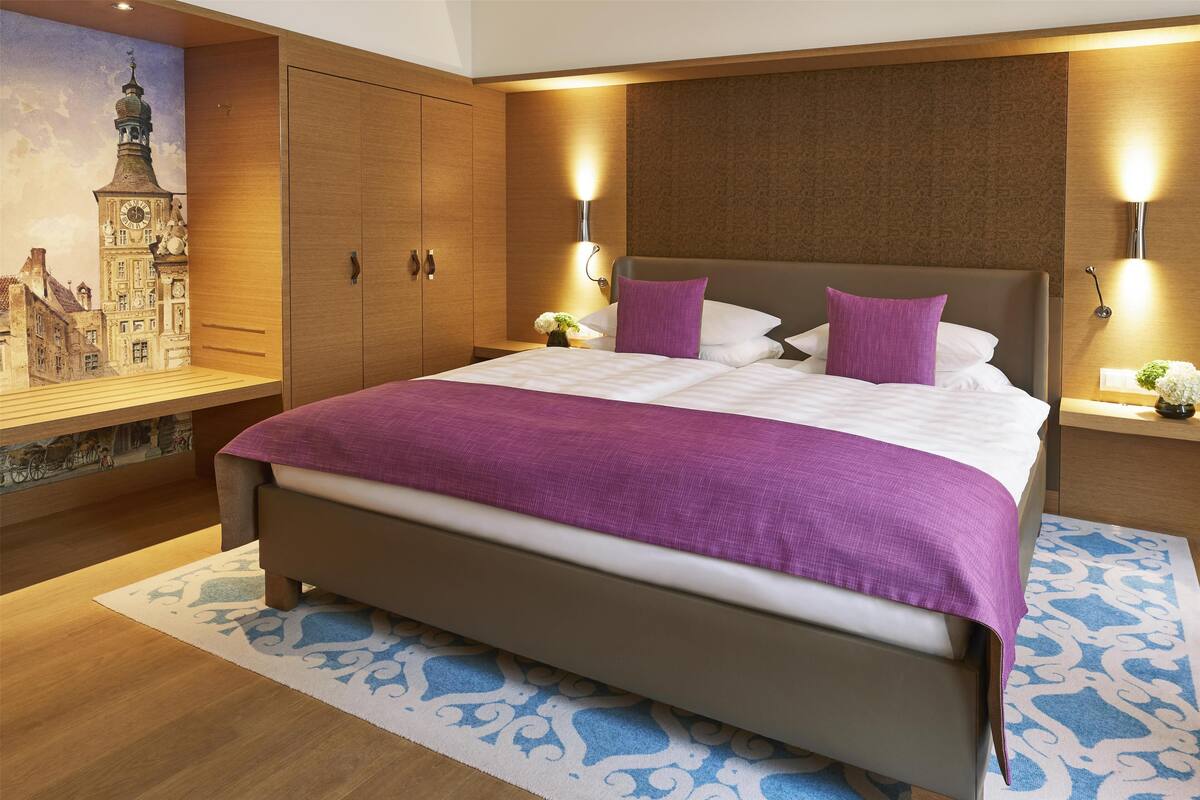 Un grand lit avec une couverture violette, une tête de lit en bois et deux lampes fixées au mur.