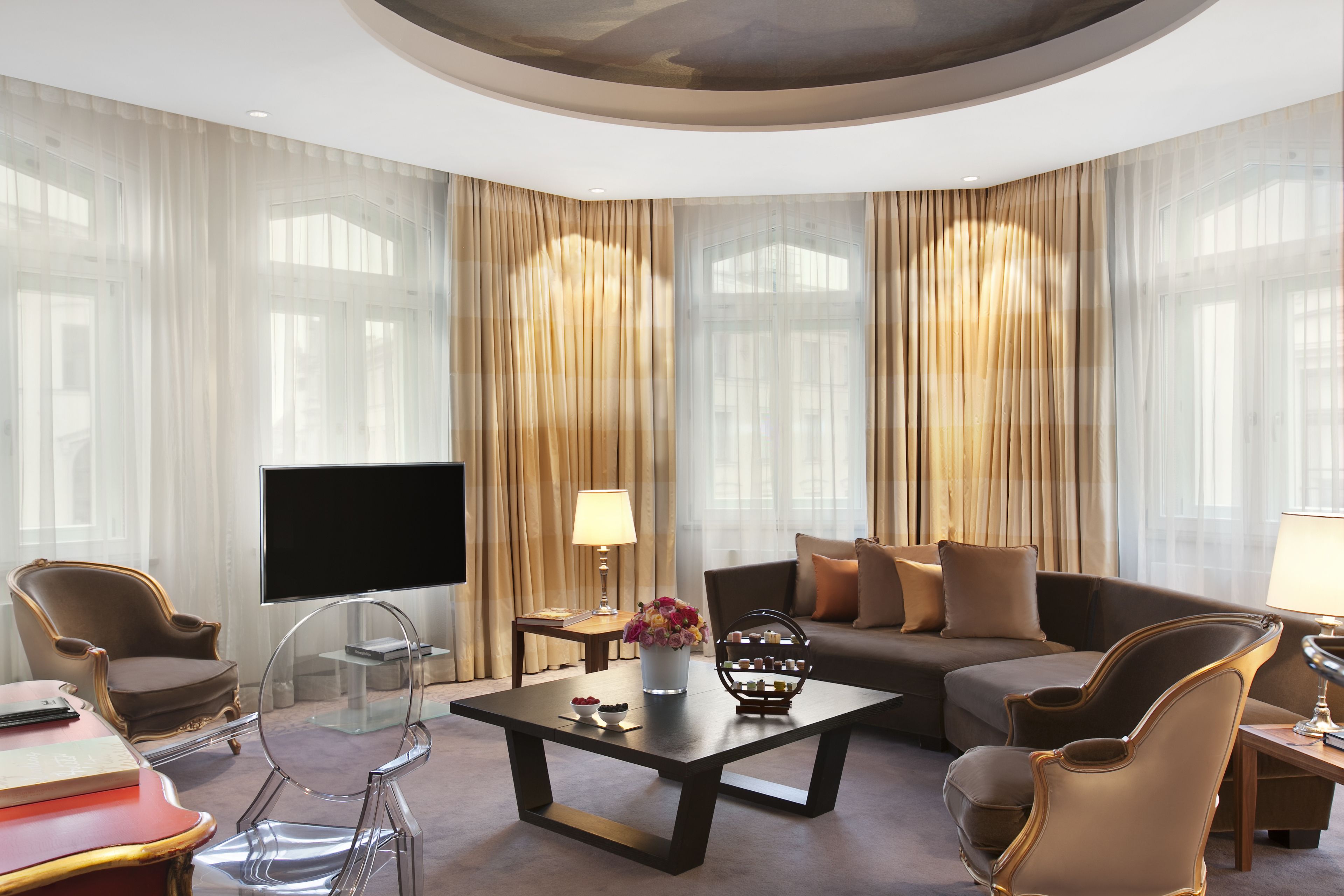 Ein modernes Hotelzimmer mit großem Flachbildfernseher, einer Couch, zwei Sesseln, einem Couchtisch und einem TV-Schrank mit Blumenvase.