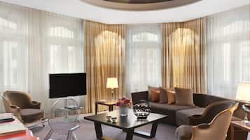 Ein modernes Hotelzimmer mit großem Flachbildfernseher, einer Couch, zwei Sesseln, einem Couchtisch und einem TV-Schrank mit Blumenvase.