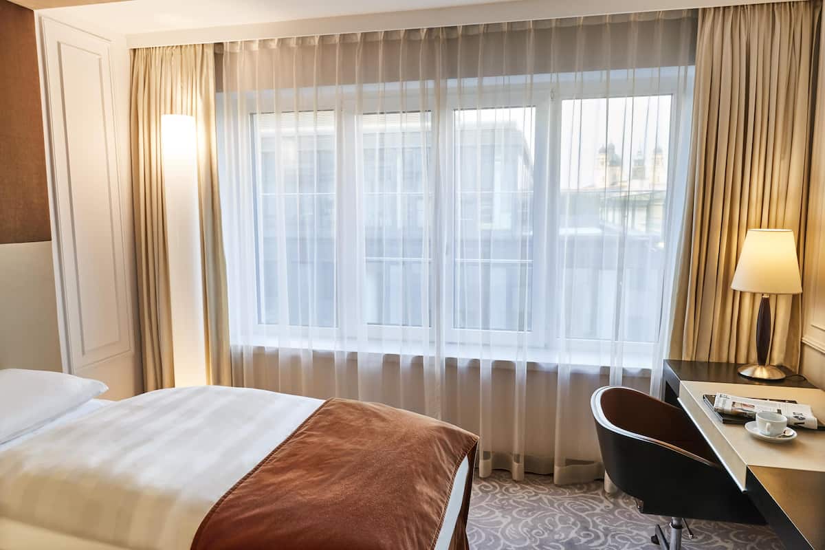 Une chambre d’hôtel avec un lit, un bureau, une chaise, une lampe et une fenêtre avec des rideaux.