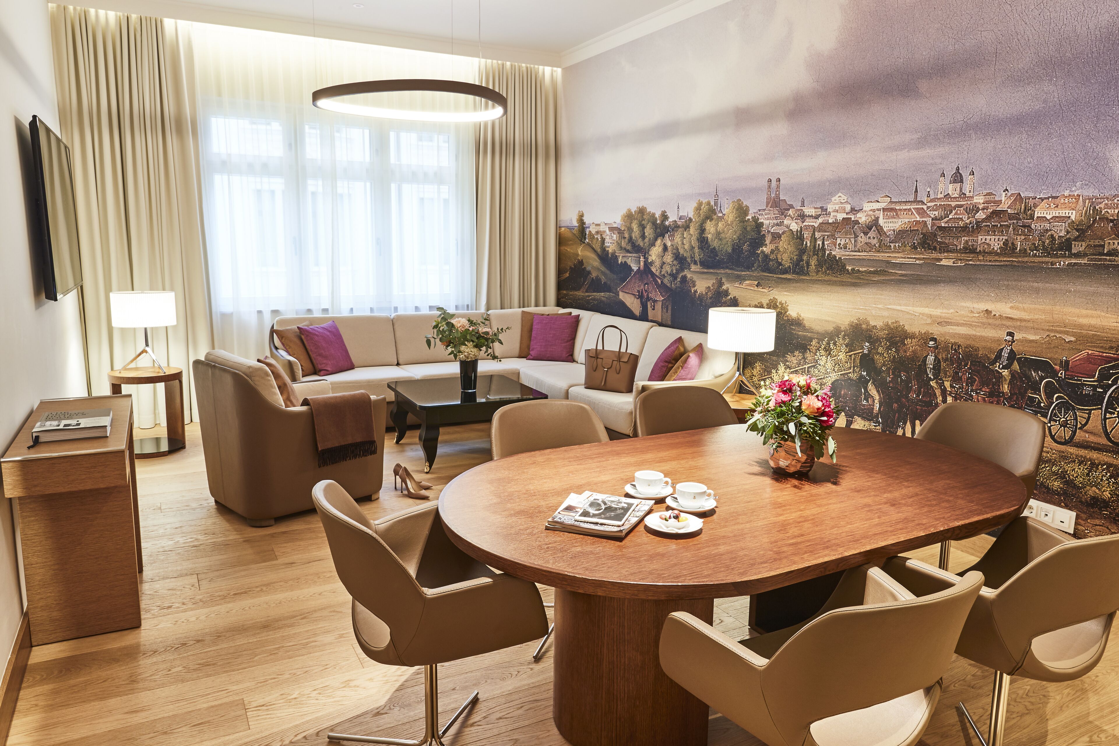 A modern hotel room with a large round dining table, comfortable seating, a sofa, and a mural of a cityscape.