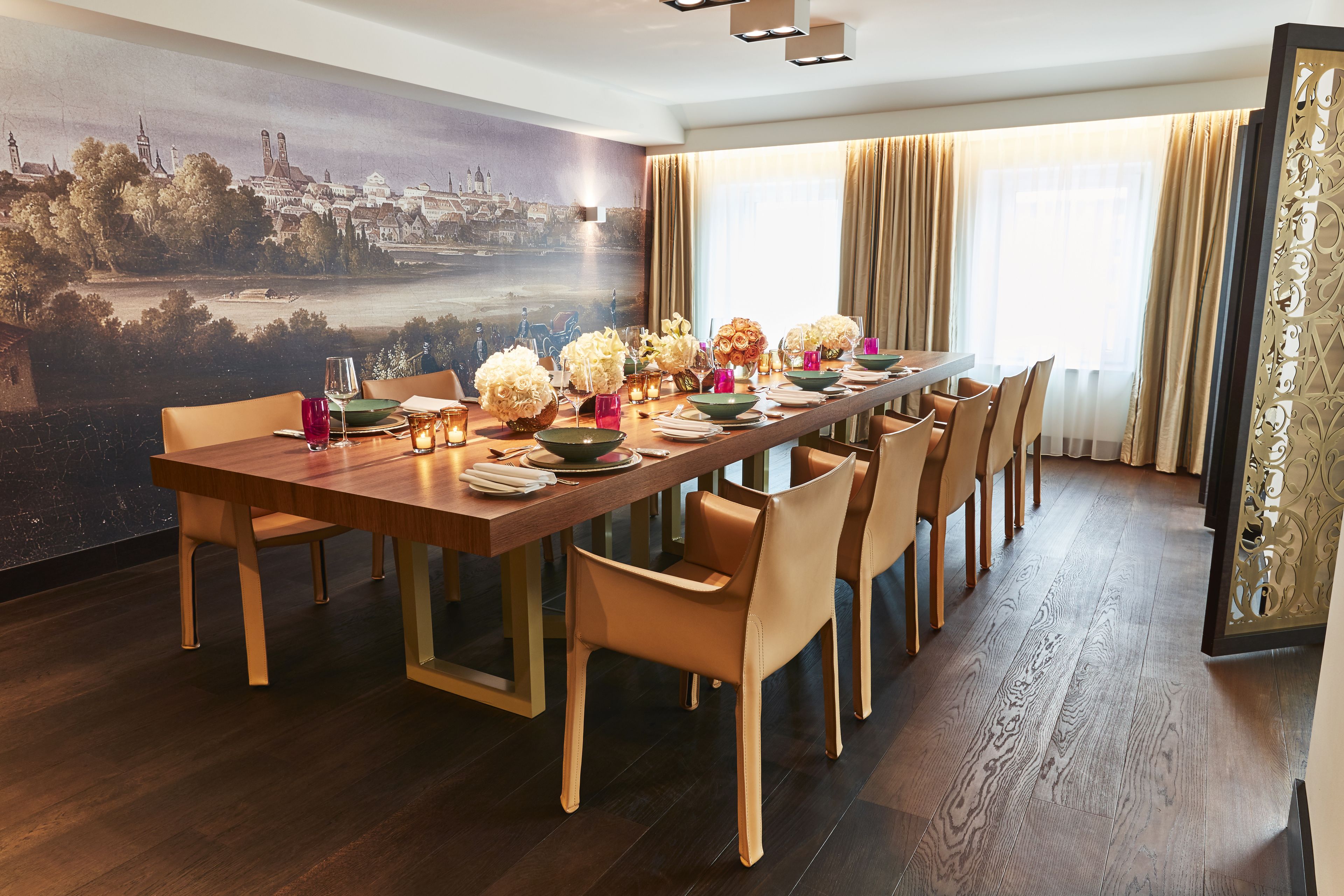 Comedor con una larga mesa dispuesta para seis comensales, rodeada de sillas, un mural con un paisaje urbano en la pared y un ventanal con cortinas.