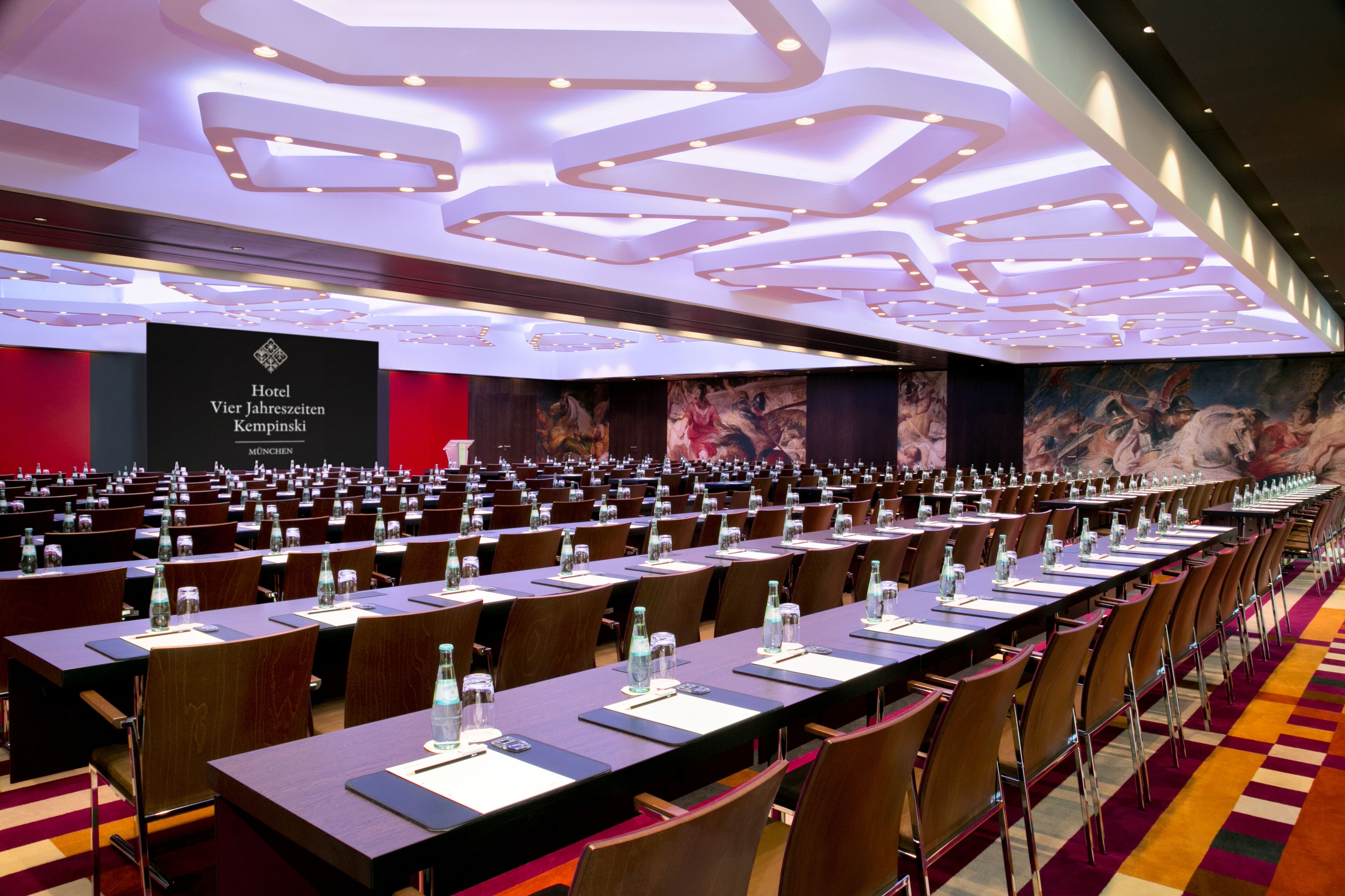 Un salón de conferencias amplio con filas de mesas y sillas, una pared roja con el letrero de un hotel e iluminación empotrada en el techo.