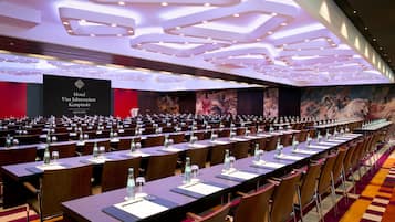 Un salón de conferencias amplio con filas de mesas y sillas, una pared roja con el letrero de un hotel e iluminación empotrada en el techo.