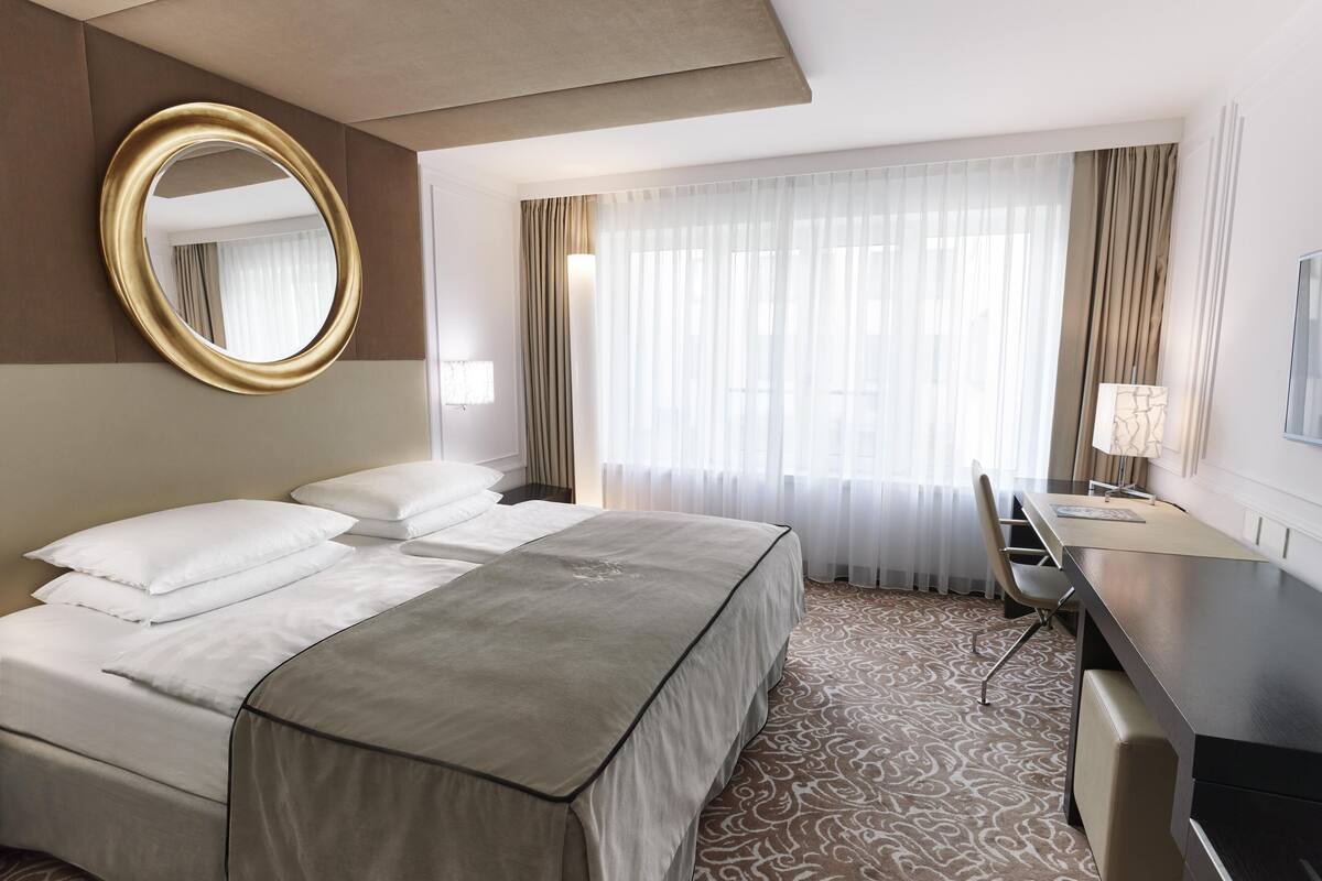 Une chambre d’hôtel avec un lit, un bureau, une chaise, un miroir rond et une grande fenêtre avec des rideaux.