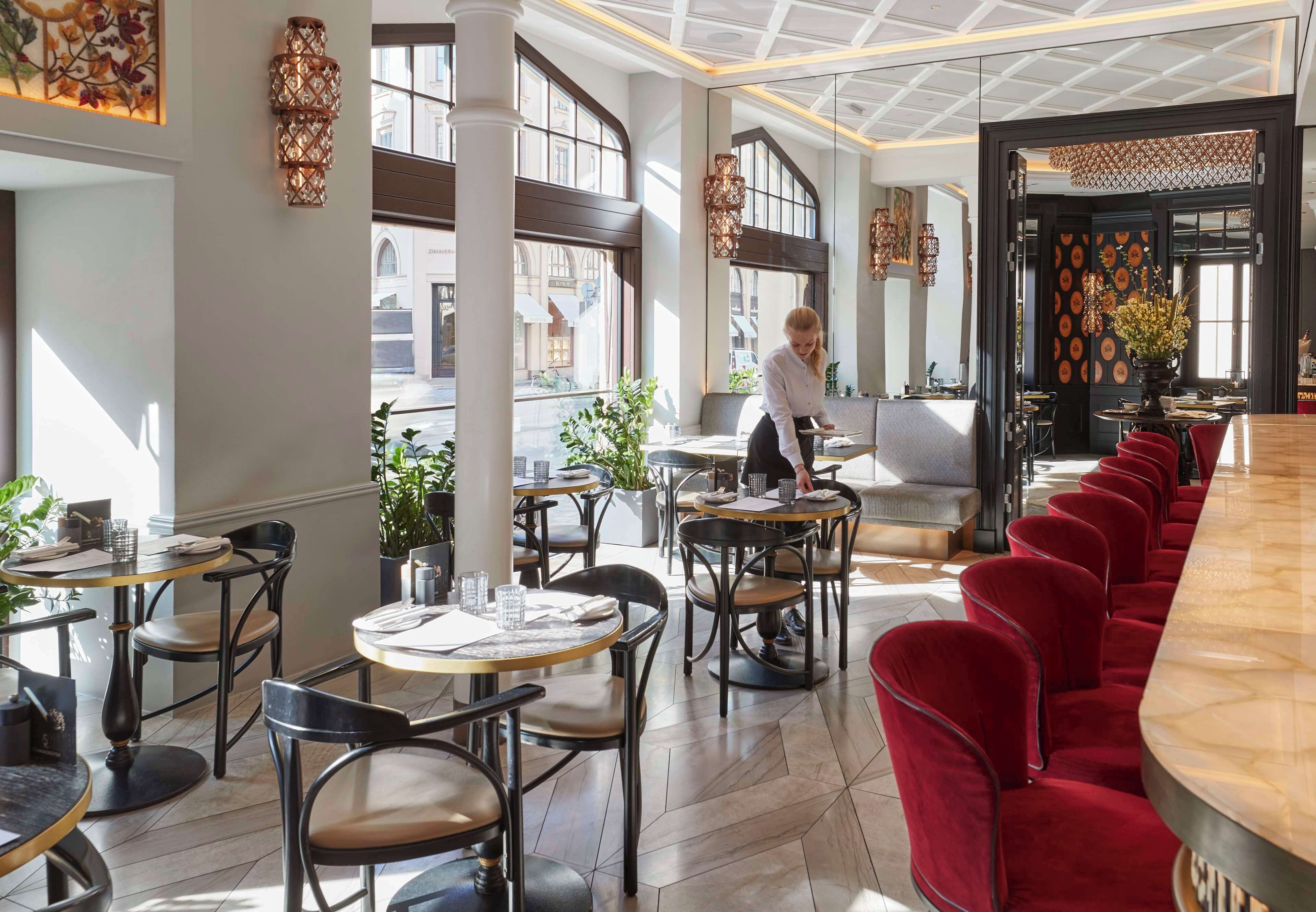 A modern restaurant with round tables and red chairs, large windows, and decorative wall sconces.