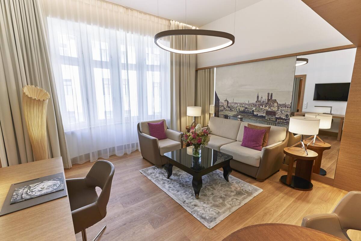 Une chambre d’hôtel avec un canapé, des fauteuils, une table basse, un téléviseur et un tableau représentant un paysage urbain.