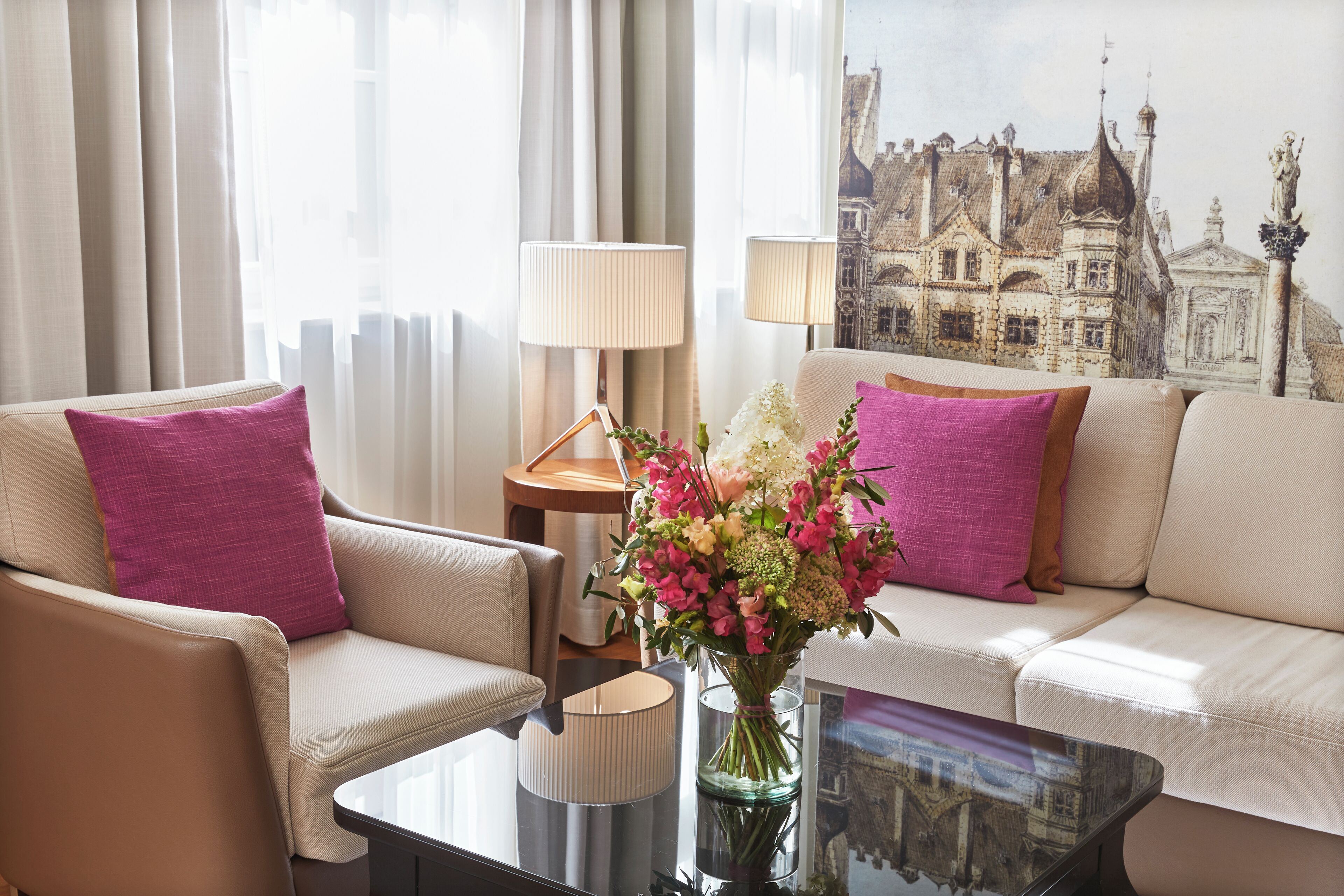 Habitación de hotel con un sofá color beige, dos sillones, una mesa de centro de vidrio y un jarrón con flores.