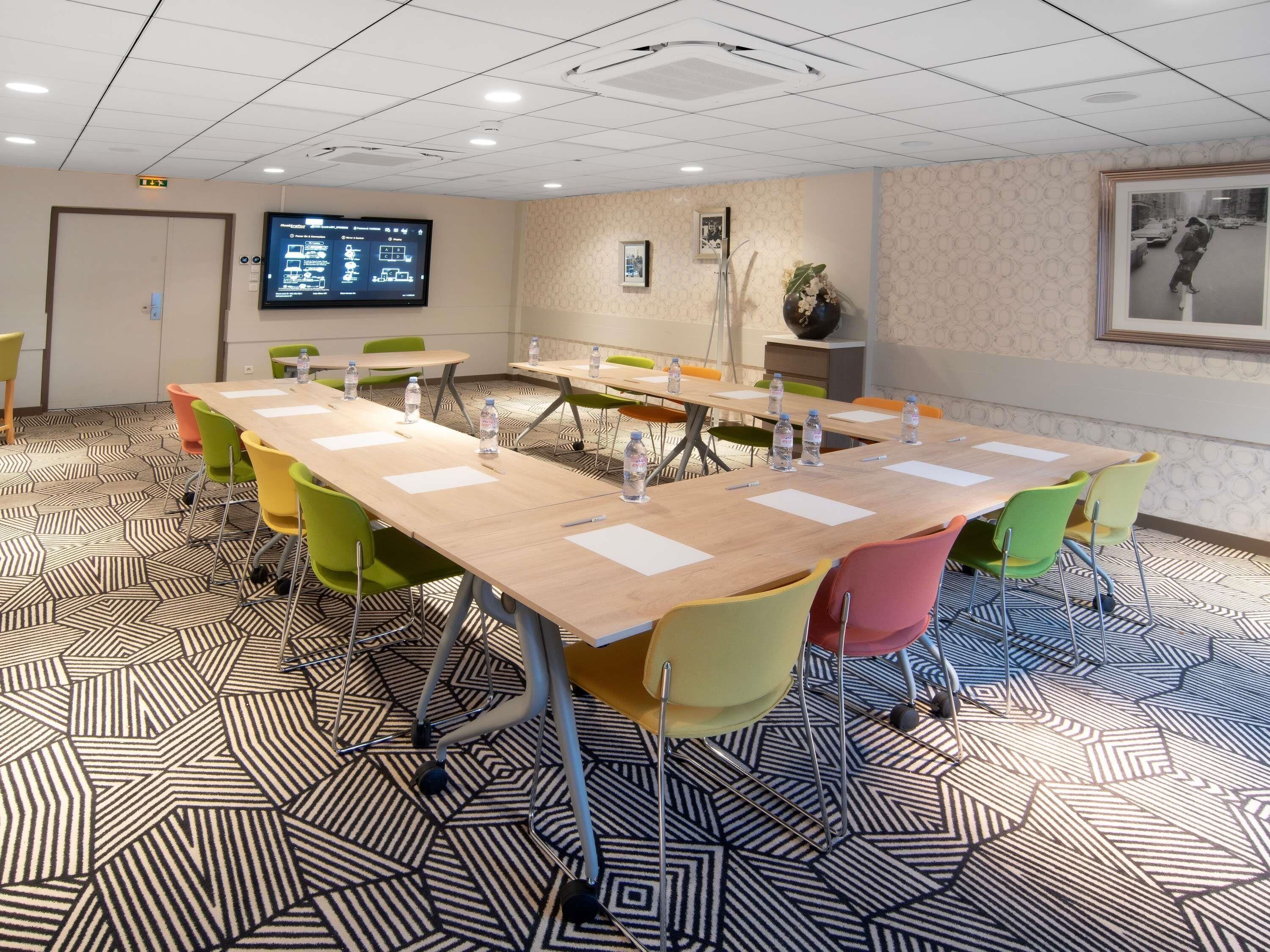A conference room with a long table, colorful chairs, and a screen displaying information.