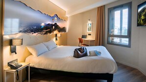 A hotel room with a bed, a desk with a laptop, a telephone, and a wall with a scenic mural. - ibis Styles Clermont Ferrand Le Brezet Aeroport (Clermont-Ferrand)