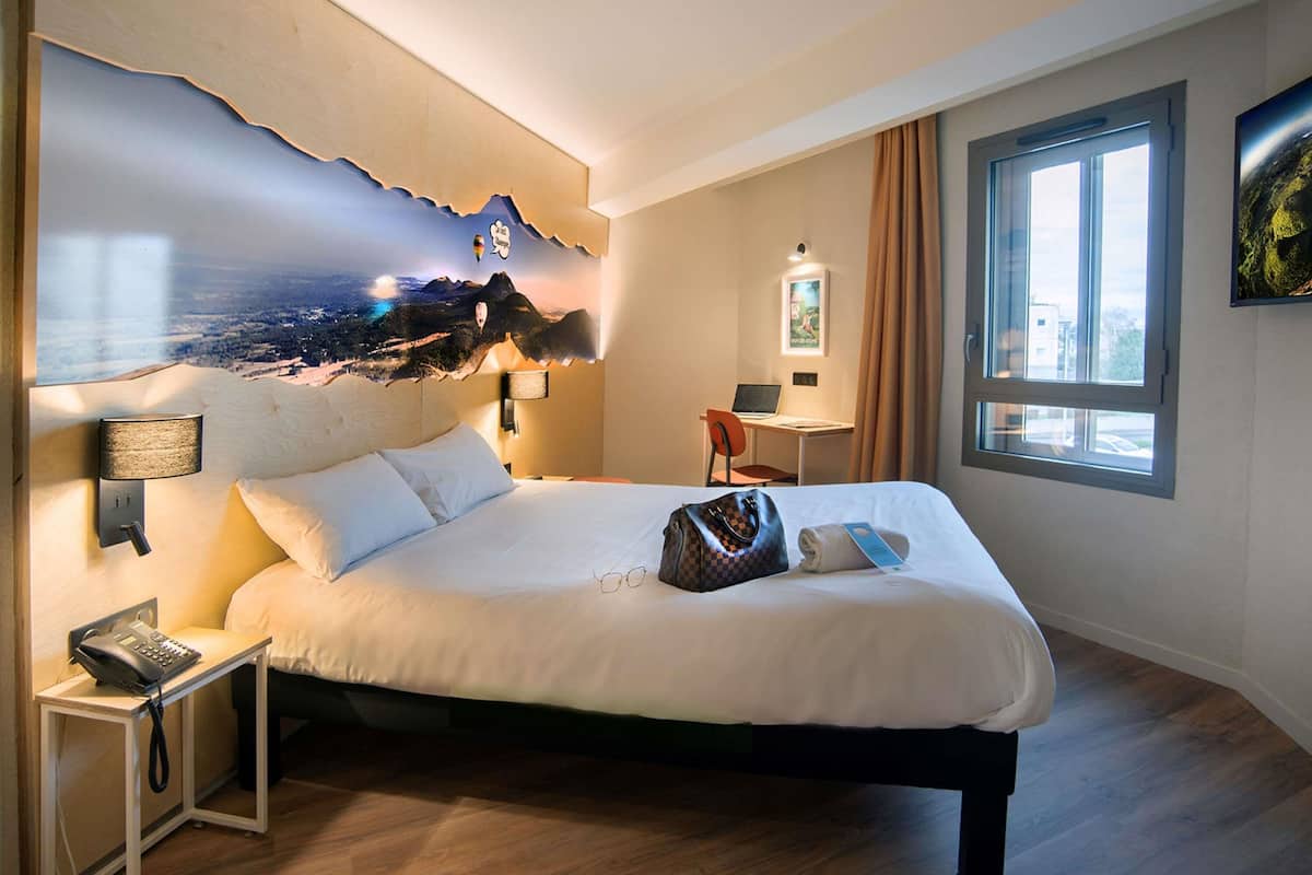 A hotel room with a bed, a desk with a laptop, a telephone, and a wall with a scenic mural.