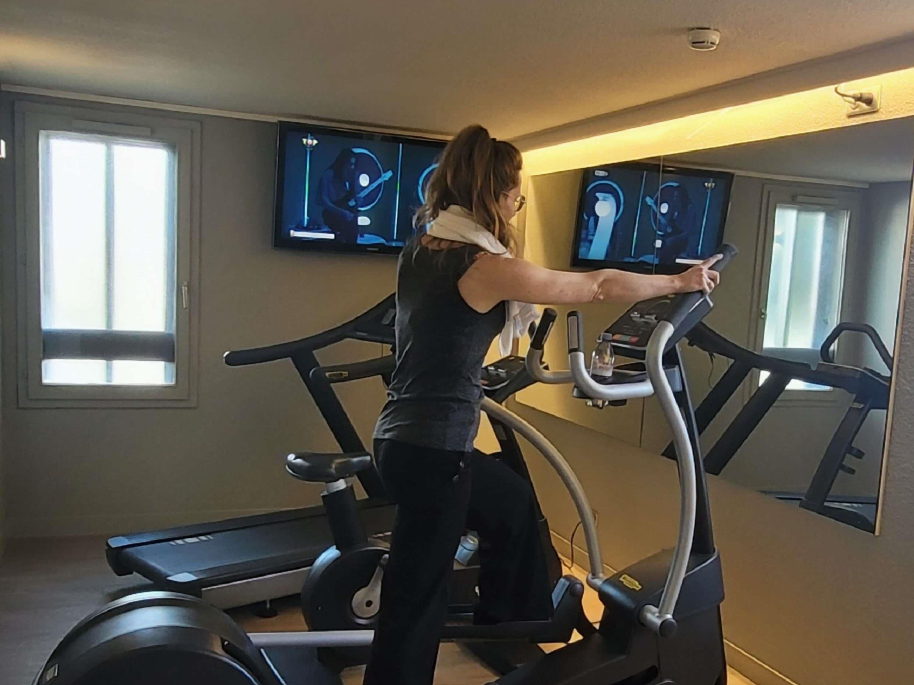 A person using an elliptical machine in a room with two windows and two televisions mounted on the wall.