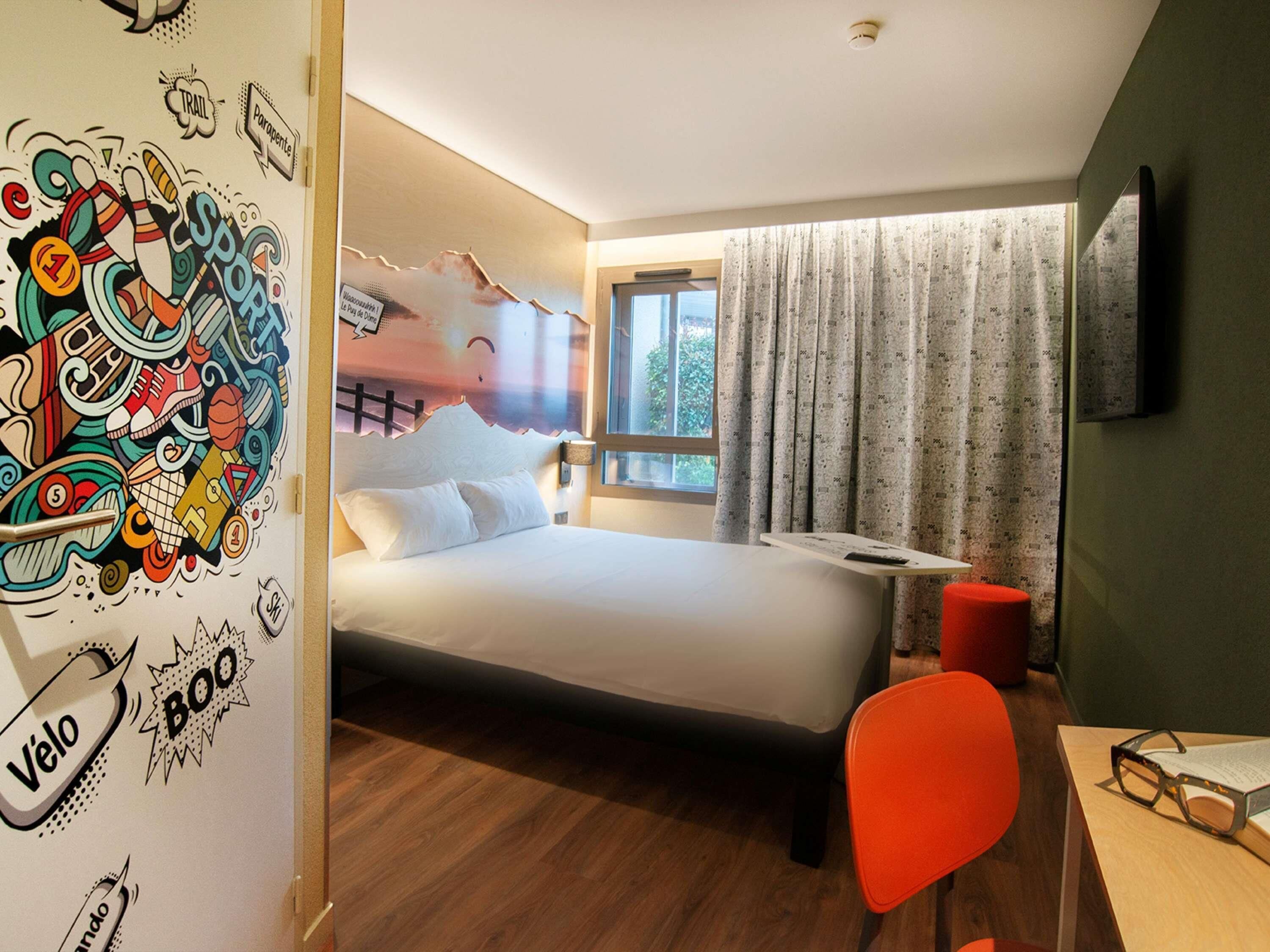 A modern hotel room with a bed, a desk, a chair, and a wall mural.