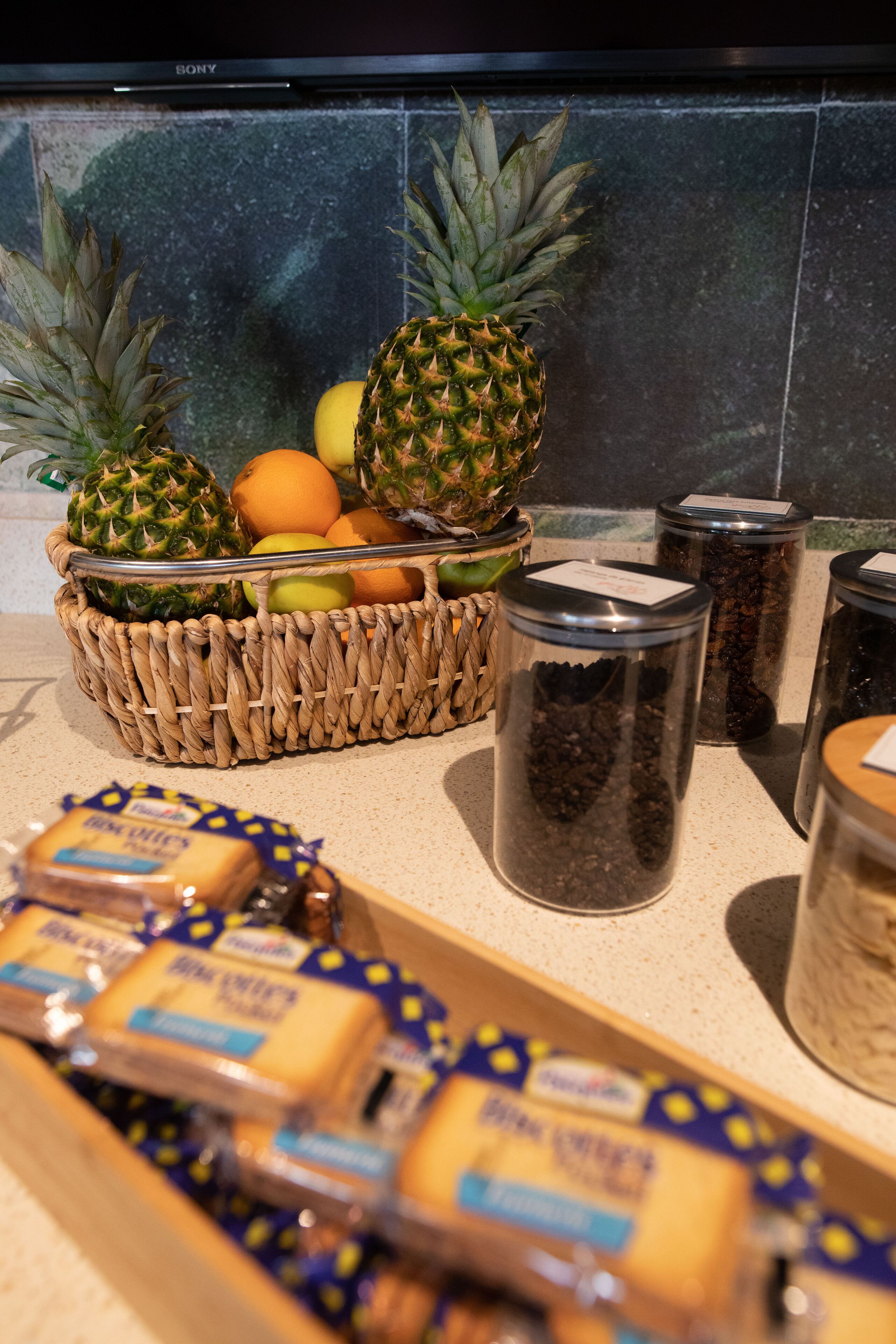 A kitchen counter with a woven basket containing pineapples, oranges, and apples, along with jars of spices and packaged food items.