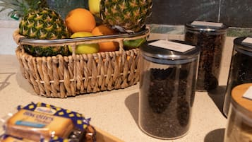 A kitchen counter with a woven basket containing pineapples, oranges, and apples, along with jars of spices and packaged food items.