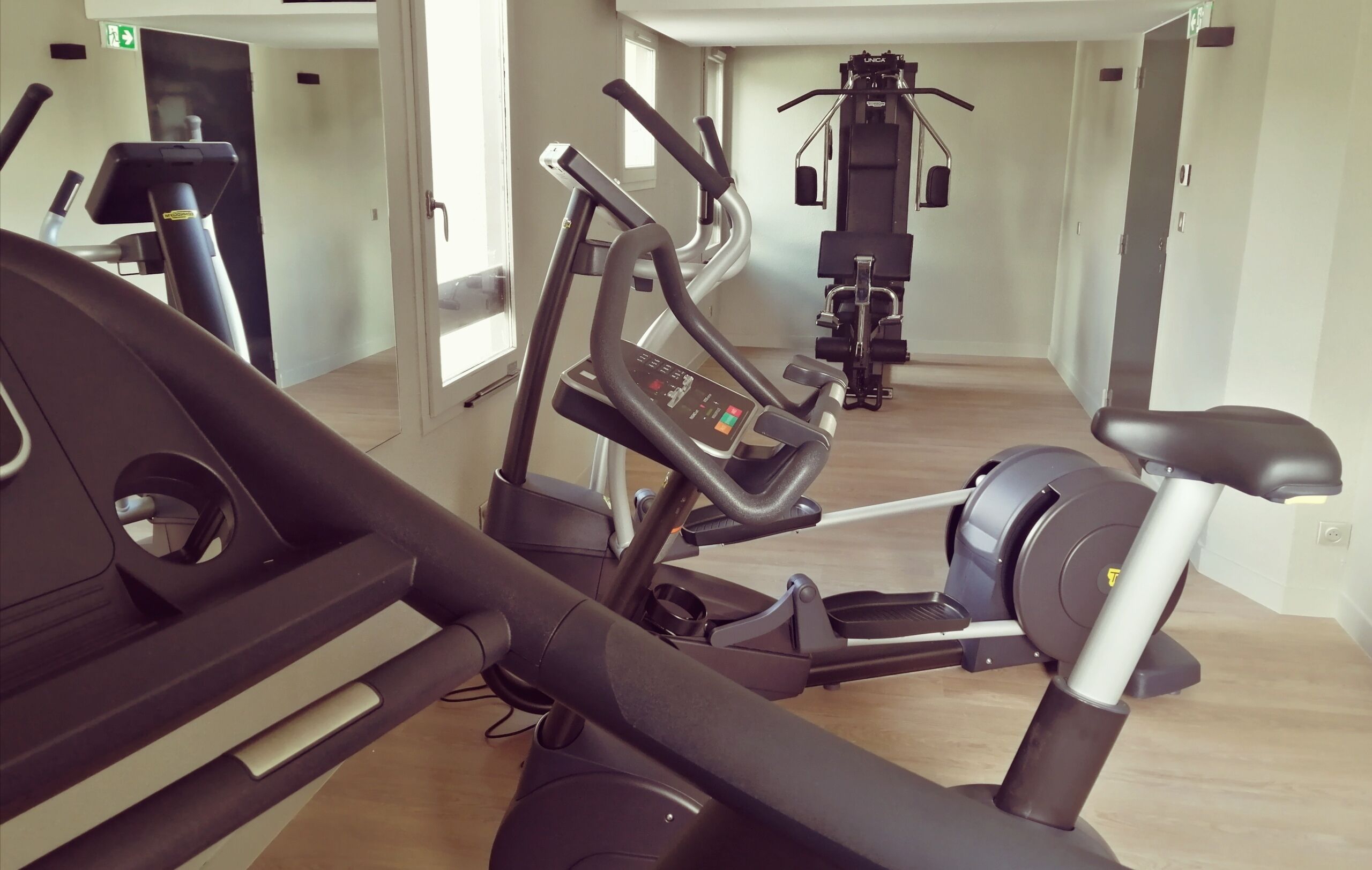 A gym with multiple exercise machines, including treadmills and elliptical trainers, arranged in a spacious room with wooden flooring and large windows.