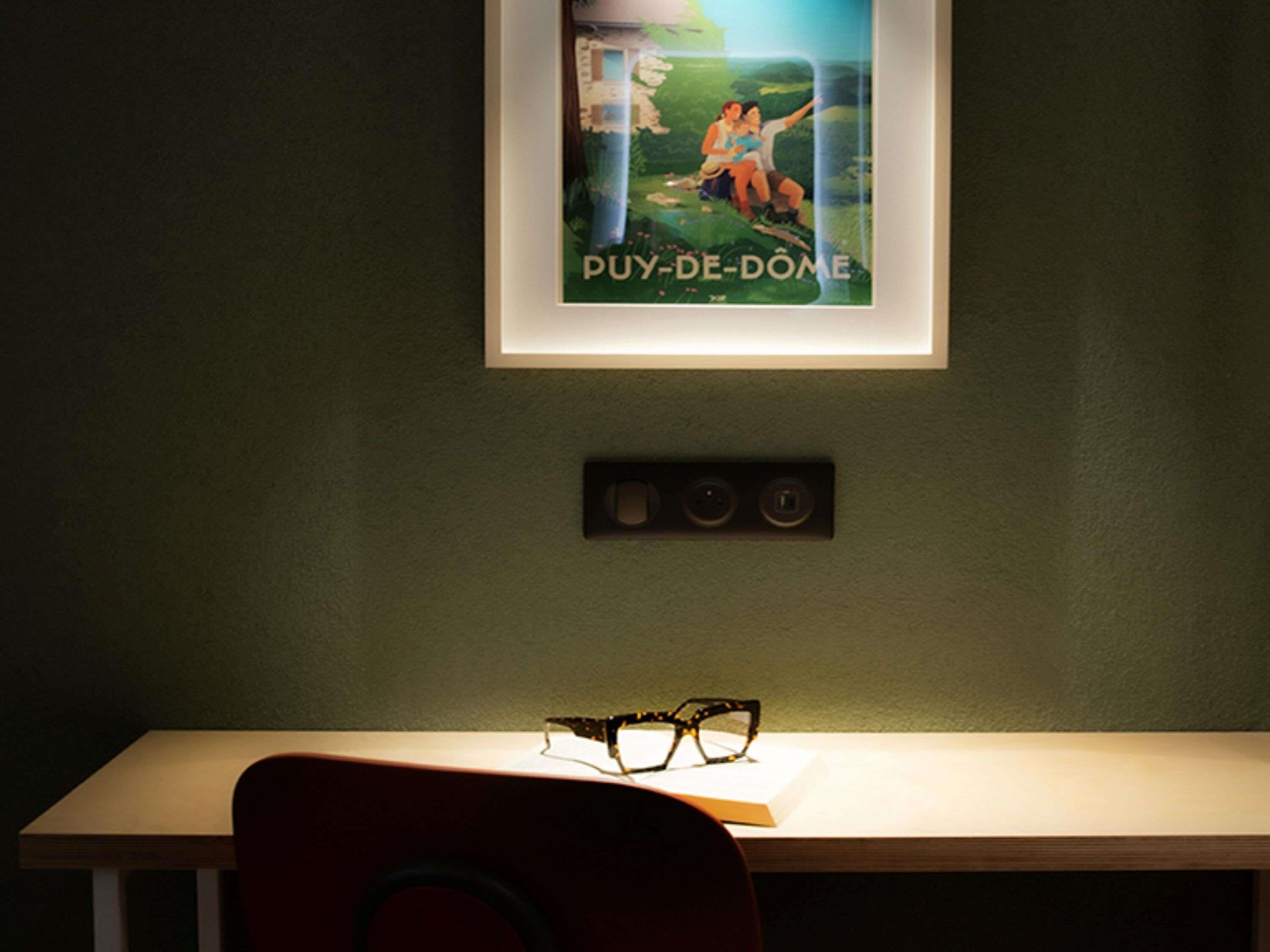 Een lijst met een poster aan de muur, een bureau met een paar brillen en een stopcontact eronder.