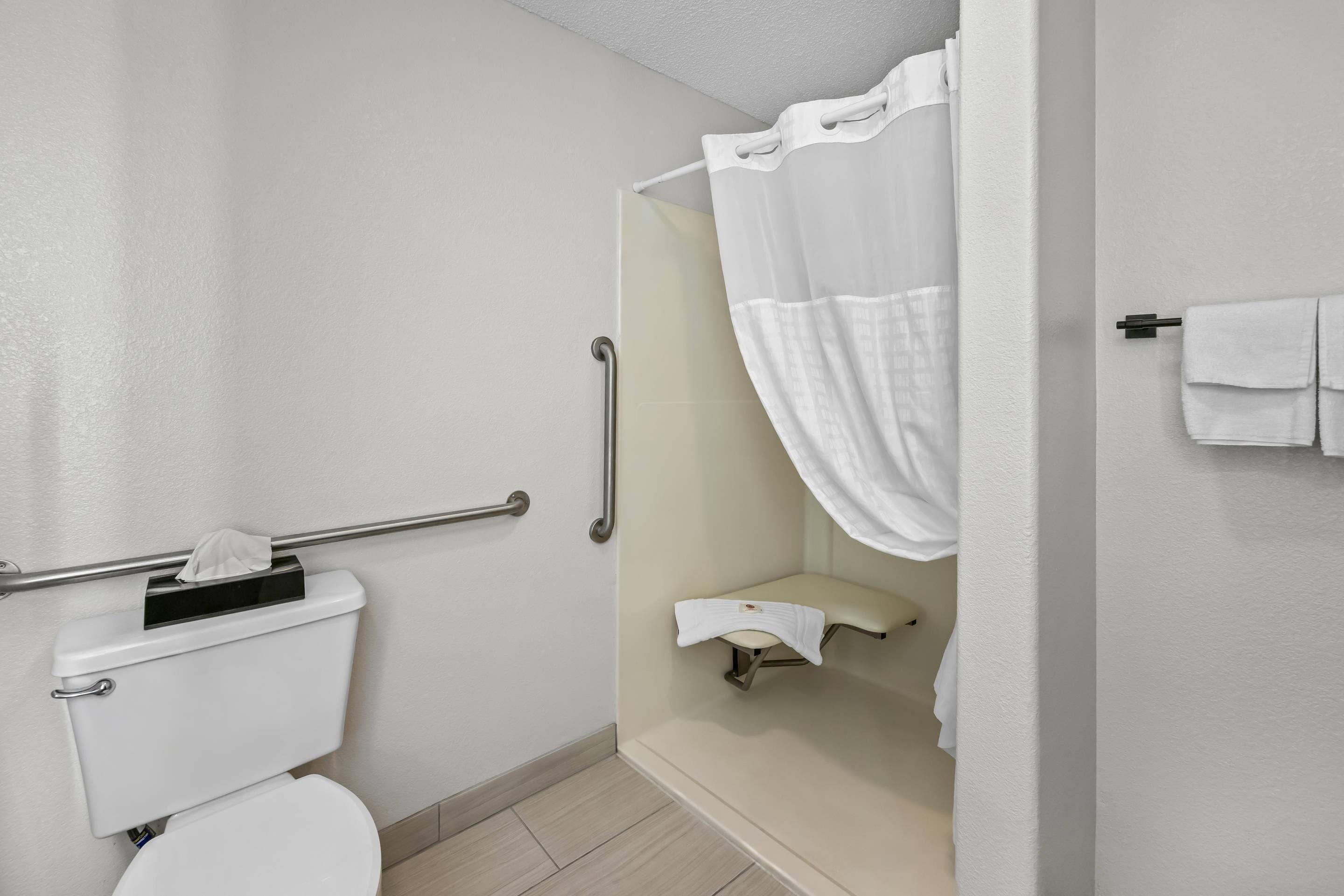 A bathroom with a white toilet, a shower with a white curtain, a grab bar, and a towel rack with towels.