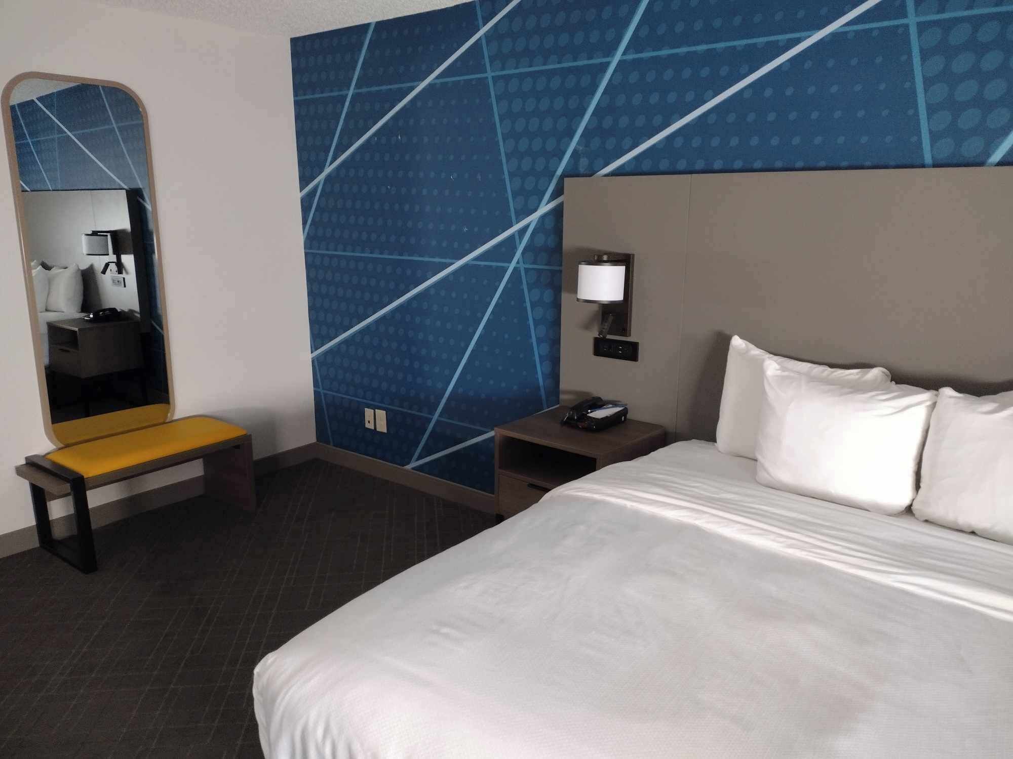 A hotel room with a large bed, a bedside table, a nightstand, a mirror, and a wall with a geometric pattern.
