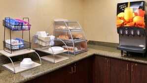 A breakfast station with a fruit juice dispenser, a display case with pastries, and a rack with paper products. - Red Roof Inn & Suites Columbus - West Broad (Columbus)