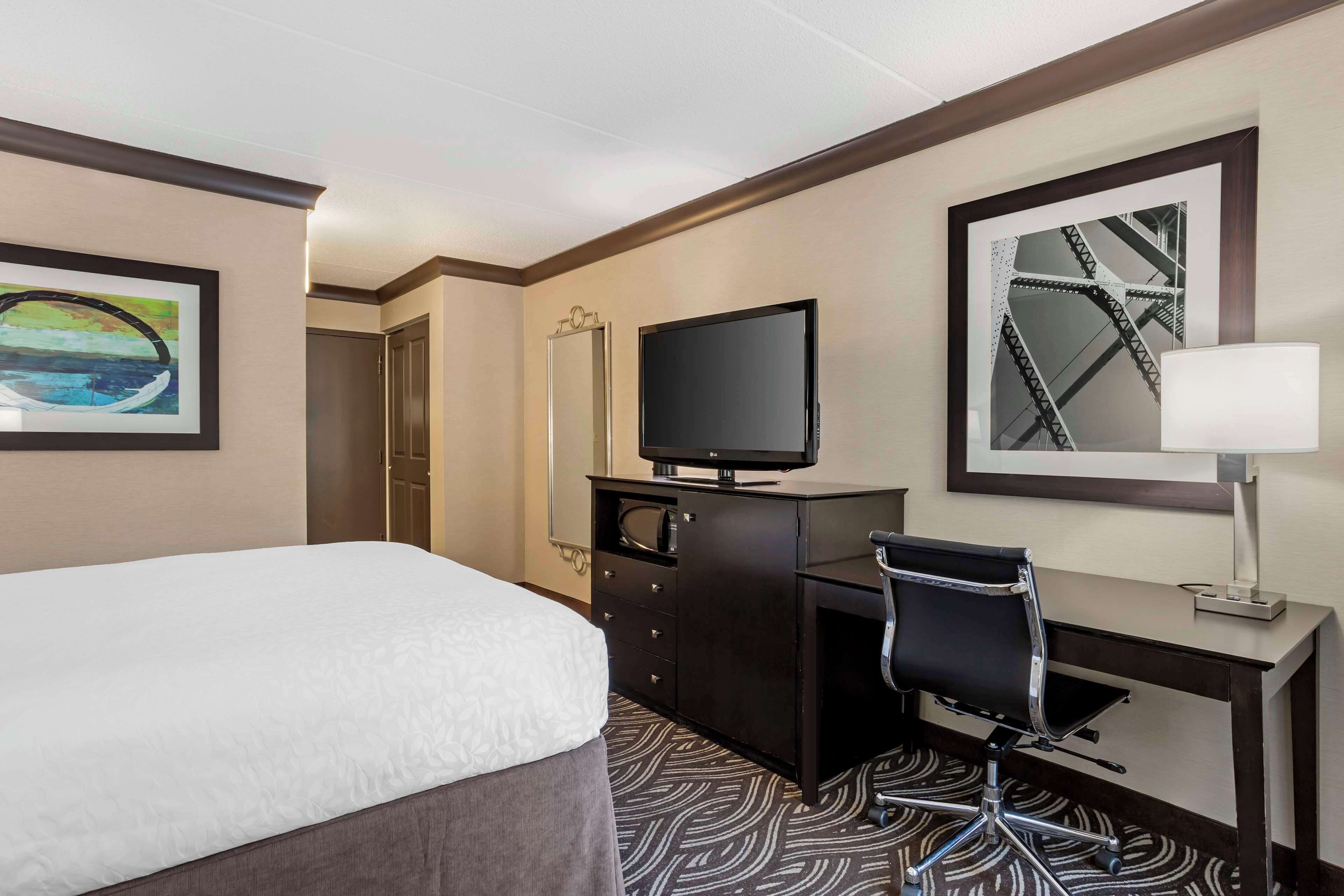 Une chambre d’hôtel comprenant un lit, un bureau avec une chaise, une télévision, un miroir et un tableau encadré accroché au mur.