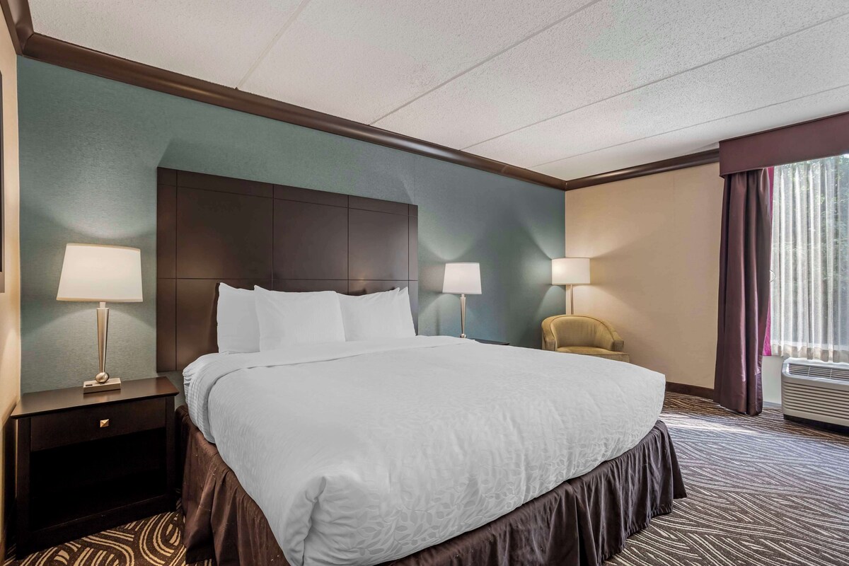 A hotel room with a large bed, two bedside lamps, a nightstand, a chair, and an air conditioning unit.
