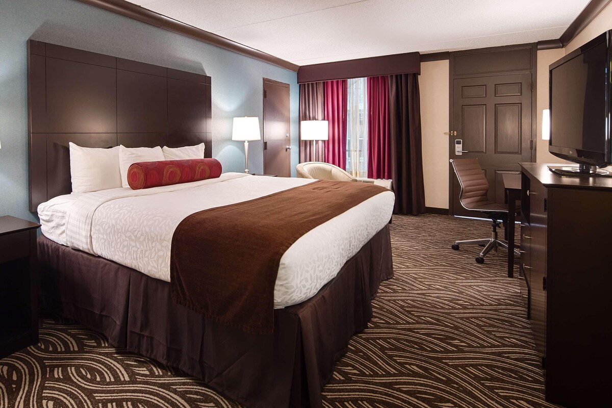 A hotel room with a large bed, a desk with a television, a chair, and a desk lamp.