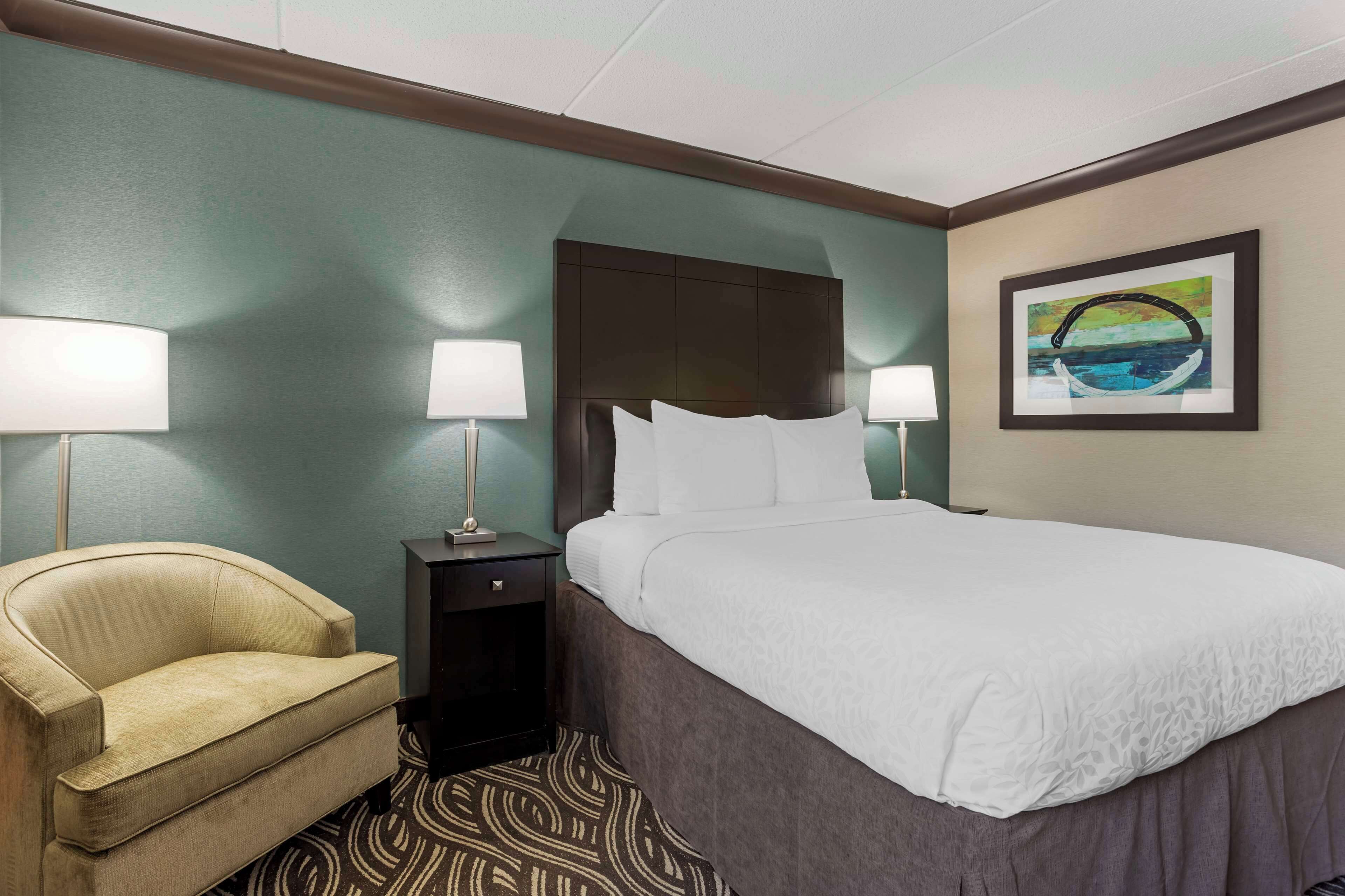 Habitación de hotel con cama, dos lámparas de noche, una silla, una mesita de noche y un cuadro en la pared.
