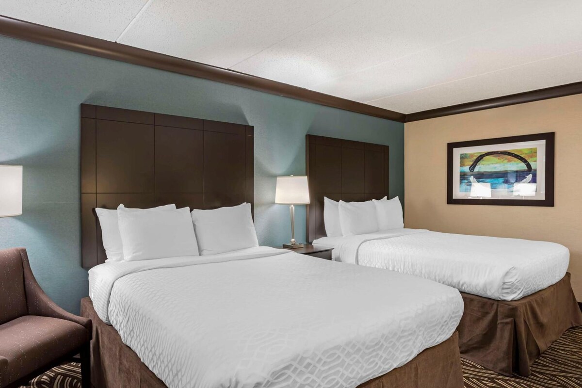 A hotel room with two beds, a headboard, a lamp, a chair, and a framed picture on the wall.