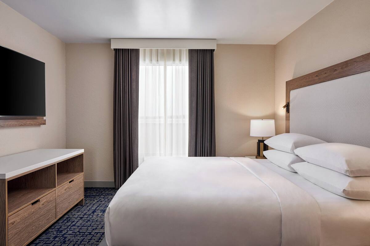 Una habitación de hotel con cama, televisor en la pared, escritorio y lámpara.