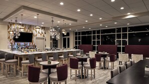 A modern bar area with a variety of seating options including bar stools and round tables, a television mounted on the wall, and a large window providing a view of the night sky. - Sheraton Richmond Airport Hotel (Sandston)