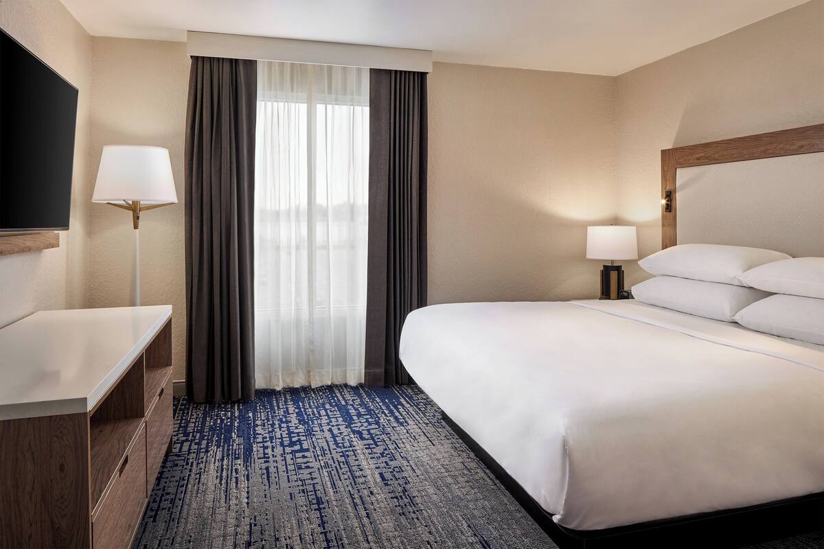 Habitación de hotel con cama, escritorio, televisor, lámpara y ventana con cortinas.
