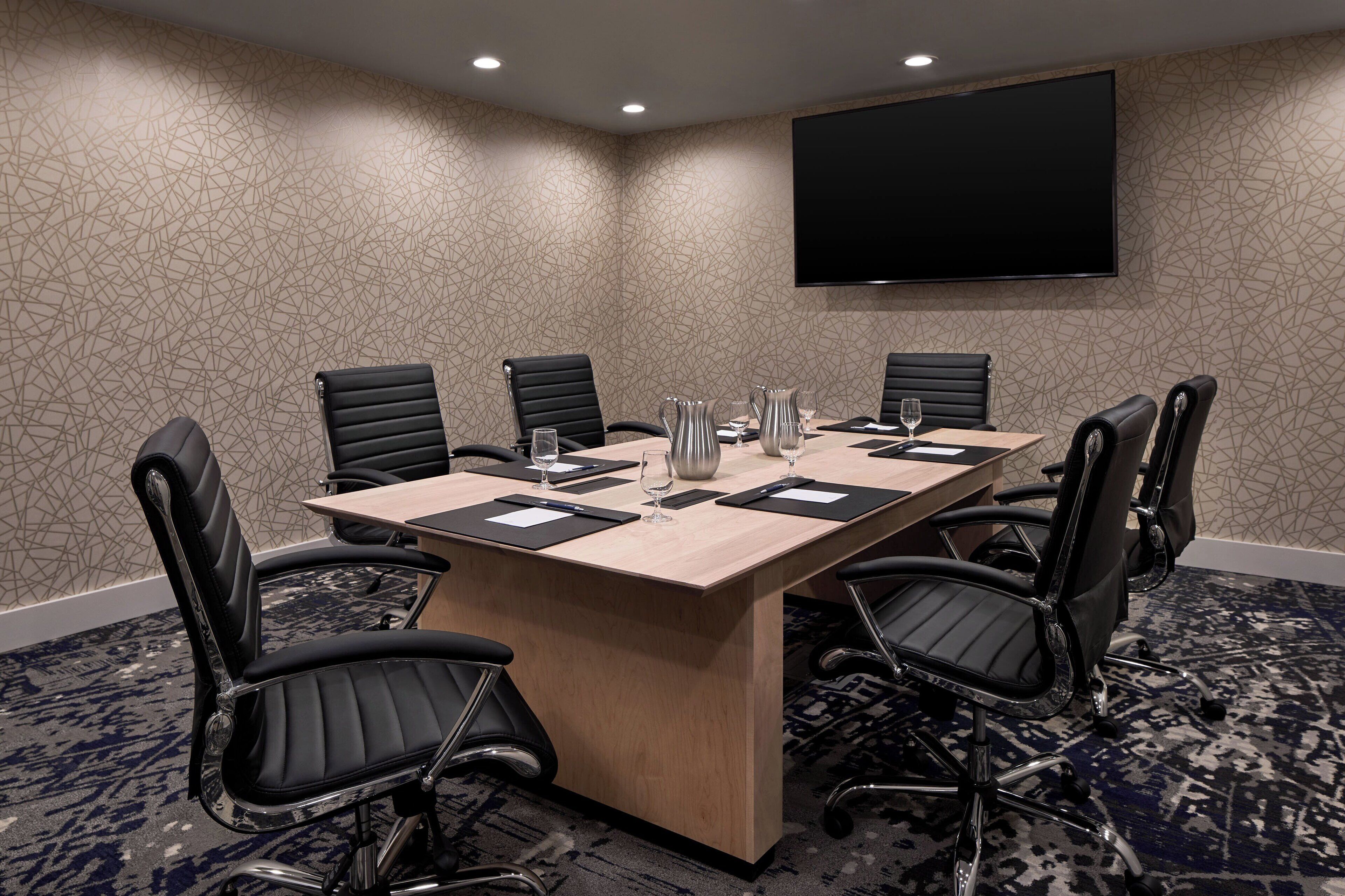 A conference room with a long table, six black chairs, a flat-screen TV, and a patterned carpet.