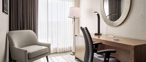 A hotel room with a wooden desk, a black office chair, a beige armchair, a round mirror, and a floor lamp.